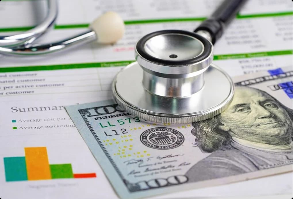 Stethoscope resting on financial spreadsheets and a hundred-dollar bill
