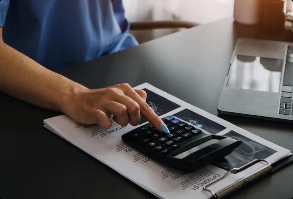 Hand using a calculator beside a laptop and paperwork