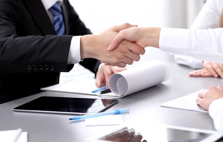 Two professionals shaking hands over a desk with documents