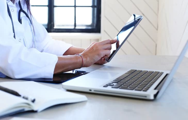 Doctor reviewing patient records on a tablet next to a laptop